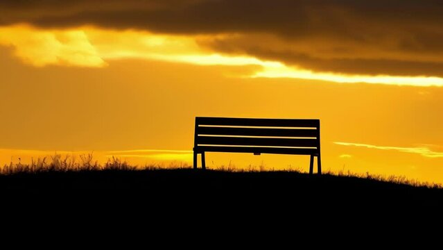 A lone bench sits atop a gry hill silhouetted against a striking golden sunset sky.