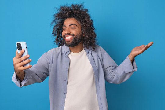 Young positive funny Arabian man takes selfie video on mobile phone camera and spreads hand with smile addressing online broadcast subscribers stands on plain blue background. Blogger, influencer