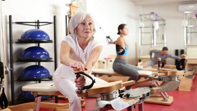 In gym, senior female performs Pilates exercises to lose weight and bring muscles of arms, chest, and body into tone using heron pulley tower. Comprehensive equipment for pumping whole body