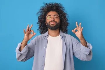 Fotobehang Young carefree smiling Arabian man with beard takes pose from yoga and zen practice to clear brain of harmful memories and gain positivity before work day stands on blue background. © antianti