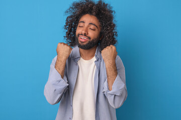 Fototapeta na wymiar Young overjoyed happy Arabian man millennial with black curly hair makes victorious wave with both hands after receiving amazing news about birth of son stands on blue background. Success, triumph