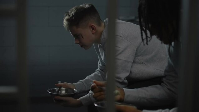 Diverse young prisoners eat disgusting prison food from iron bowls sitting on bed in prison cell. Teenage criminals serve term for crimes in jail. Juvenile detention center or correctional facility.