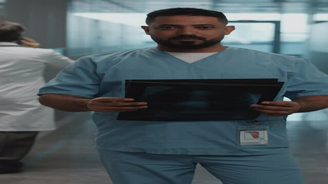 Vertical portrait of middle eastern physician in medical uniform checking x-ray images and posing on camera in hospital corridor