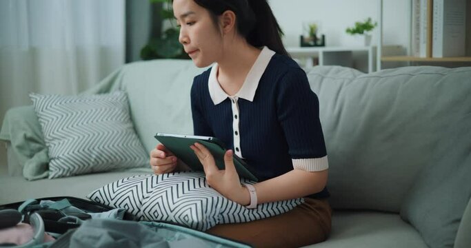 Footage slow motion shot, Asian teenager woman sitting on sofa using digital tablet making check list of things to pack for travel, Preparation travel suitcase at home.