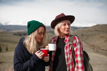 Two traveler girls with mugs stand on rock in mountains - 766807540