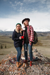 Two traveler girls with mugs stand on rock in mountains - 766807565