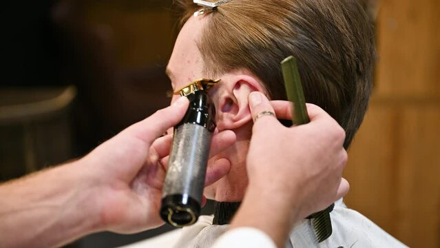 Close-up photo of professional barber shaving sideburns. Holding ear to make straight line. Competent hairdresser using electric shaver for work.