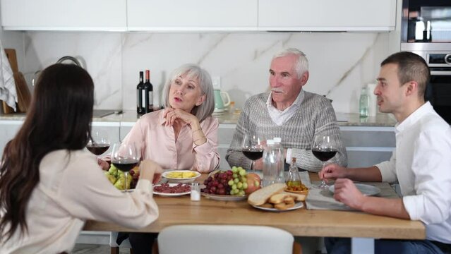 Mature spouses sitting at table, enjoying food and wine, chatting talking laughing with adult son and his girlfriend. In homely atmosphere male and female pensioner celebrate anniversary with guests