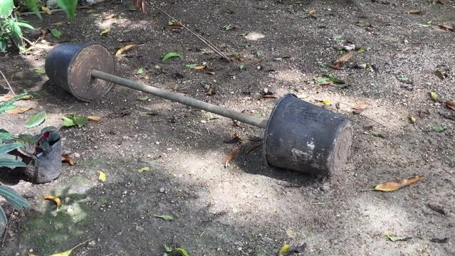 A homemade weightlifting bar on the ground in the park. The weights are made of two flowerpots filled with cement. There are leaves on the ground from the trees.