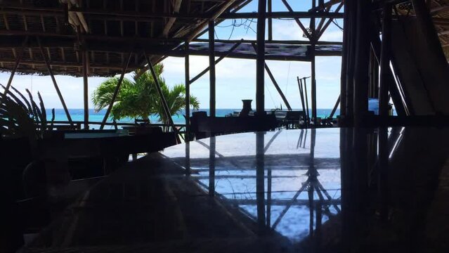 The view outside of the turquoise water on the beach from the restaurant in Siquijor Island. There are plants outside the open windows. The bar counter is dark with reflections from the ceiling.