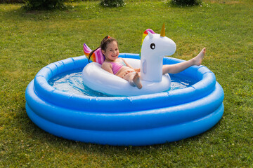 A little girl swims in an inflatable pool with a unicorn toy on a summer day in the garden.