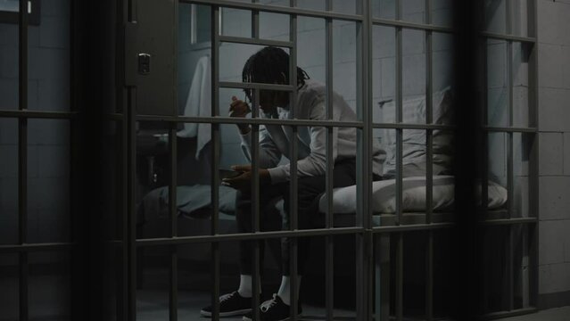 African American young prisoner eats disgusting prison food from iron bowl sitting on bed in prison cell. Teenage criminal, inmate serves imprisonment term for crime in jail. Youth detention center.
