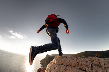 Active hiker with backpack jumps to big rock against sunset sky and sea