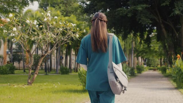 Back view of female nurse in medical uniform going home through park after shift in clinic