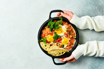 Hands hold a frying pan with Shakshuka. Poached eggs in a spicy tomato pepper sauce. Traditional Jewish scrambled eggs. Top view, flat lay.
