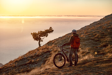 Sporty man athlete is standing with mtb bike at mountaintop and enjoys sunset or sunrise - Powered by Adobe