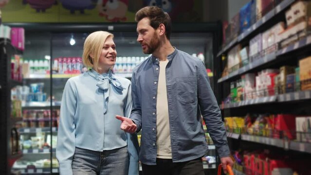 Lovely marriage couple engaged in conversation in grocery store near shelves stocked with various products. Attractive caucasian family doing shopping together in romantic atmosphere indoors.