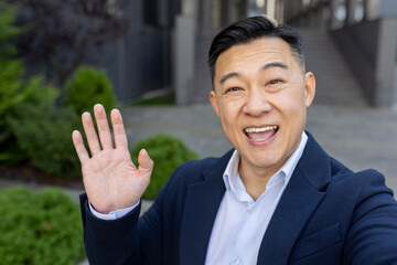 Close-up photo of a young Asian male businessman standing outside a building in a suit, talking on a video call on the phone, greeting and waving at the camera