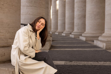 giant white columns and road between.woman female posing, in long coat. girl sitting in sunlight and shadow dropped on asphalt. architectural antic temple.