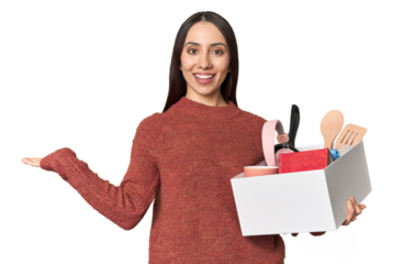 Caucasian young woman holding a box for moving on studio background showing a copy space on a palm and holding another hand on waist.