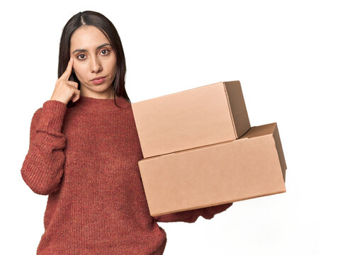 Caucasian young woman with moving boxes on studio background pointing temple with finger, thinking, focused on a task.