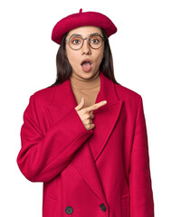 Stylish young Caucasian woman in red trench coat and beret on studio background pointing to the side