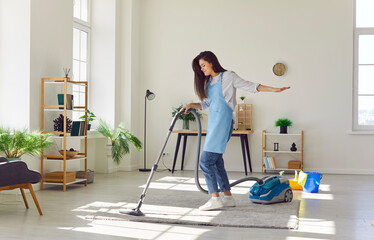 Young housewife having fun while cleaning up at home. Happy beautiful woman in apron vacuuming rug...