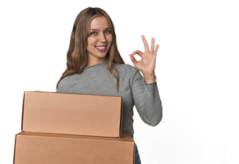 Blonde woman with cardboard boxes moving on studio backdrop cheerful and confident showing ok gesture.