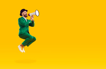 Fototapeta na wymiar Man in green suit shouts into loudspeaker announcing crazy discounts for St. Patrick's Day. Young stylish man jumping high with megaphone on orange background. Copy space. Full length. Web banner.