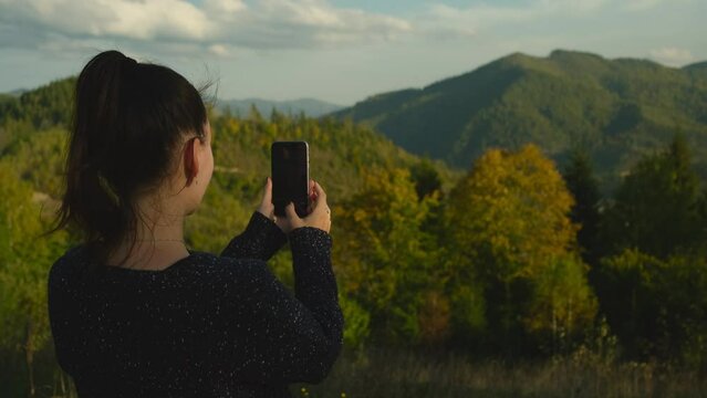 Young woman making photos of mountains on cellphone camera. Happy girl standing on top of mountain peak and photographing scenic landscape using mobile phone device