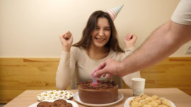 A girl sits in front of a table with a festive cake, into which a candle in the form of the number 16 is stuck. The concept of a birthday celebration.