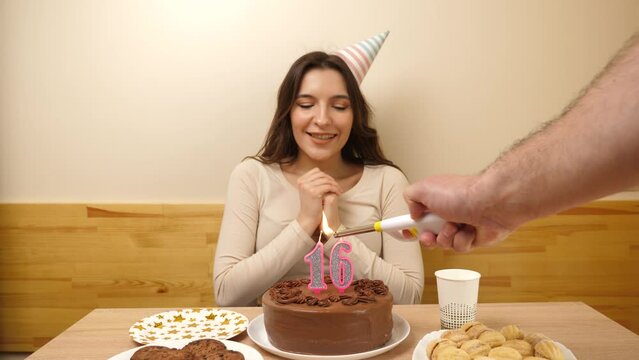 The girl is sitting in front of a table with a festive cake, in which a candle in the form of the number 16 is burning, which she blows out. Birthday celebration concept.