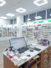 Blurred view inside of pharmacy with the seller counter, computer and cash register against stocked shelves with medicines. Healthcare and cosmetics industry defocus background