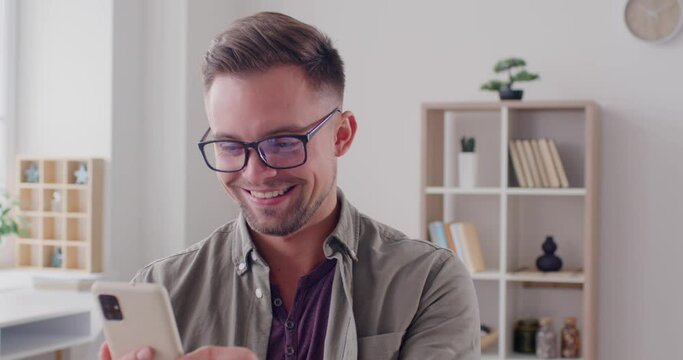 Happy cheerful man scrolling social media on phone. Smiling young man in good mood using cellphone, browsing entertaining photo video content, laughing at funny reels, enjoying virtual communication