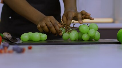 The cook collects grapes from a bunch to prepare a fruit salad, the cook peels a bunch of grapes with his hands for a fruit salad, and grapes are prepared for a fruit salad on the kitchen table.