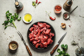 Raw diced beef meat on circle cutting board with fork and knife, herbs and spices for tasty cooking. Goulash food preparation. Top view