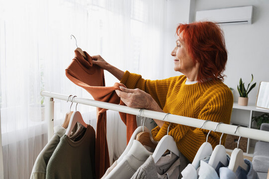 Retired redhead senior woman choosing sweater from clothes rack at home