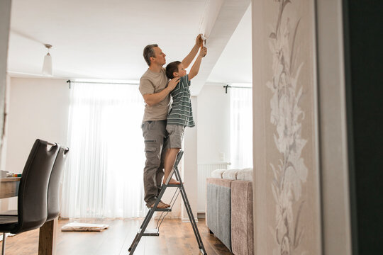 Son and father peeling old wallpaper standing on step ladder at home