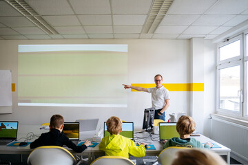 Professor pointing at projection screen and teaching computer to boys in classroom