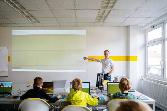 Professor pointing at projection screen and teaching computer to boys in classroom
