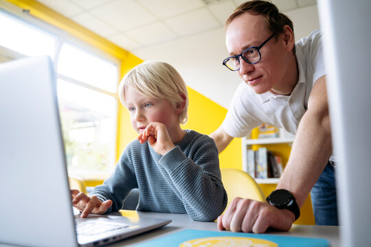 Blond boy using laptop by professor leaning on desk in classroom