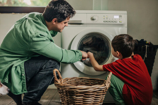 Father and son loading clothes from basket in washing machine at home