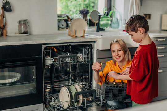 Mother and son holding dirty spoons near dishwasher in kitchen at home