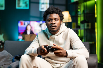 Bored person plays online videogame, spamming attack buttons on gamepad. Gamer in neon lit apartment participating in internet esports competition, sitting on couch and playing on gaming console - obrazy, fototapety, plakaty