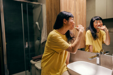 An asian woman is brushing teeth in a bathroom.