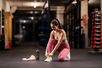 Fit sportswoman is kneeling and tying her shoelace in a gym.