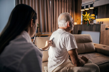 Rear view of a senior man sitting on couch at home while a doctor is examining lungs.