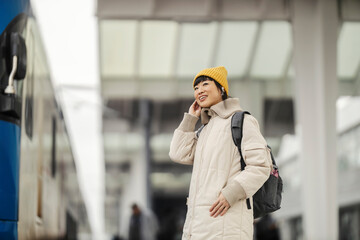 A japanese passenger is standing at train station, waiting for a train and putting earphones in ears.