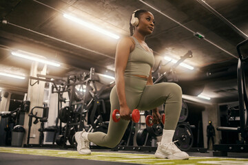 A fit young black sportswoman practicing lunges with dumbbells and listening music on headphones at gym.
