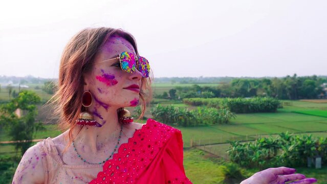 Caucasian young woman celebrating Holi face covered with colorful paint pink purple wearing sunglasses outside. Holi festival of colors. India, traditions, jewelry, body art, artistic make up